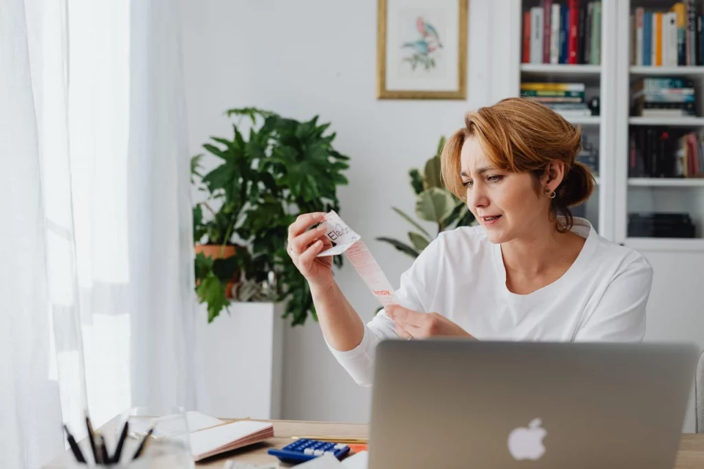 karolina-grabowska-5900134 Carbon Accounting being carried out by young woman in brown hair and white top holding a paper while sitting with an apple laptop
