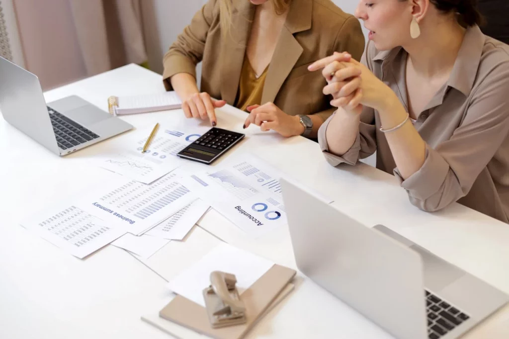 chase strategy being analysed and prepared to be used on a project where 2 women are discussing using data mentioned on papers kept on a white table next to a laptop