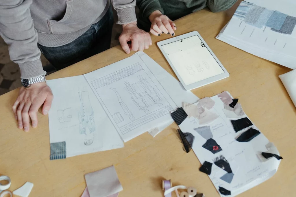 pexels-cottonbro-4622224 Papers and drawings layed on a wooden table with cloth sample stapled to them in some and rest with drawings of figures in fashion and on an iPad as well. We build continuous improvement with our actions in daily decisions in our business