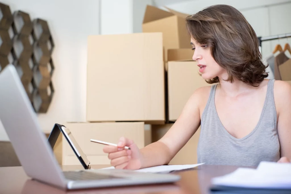 A woman wearing grey top and short brown hair managing Operational Inefficiencies looking at a laptop while having cardboard boxes stacked up in the background