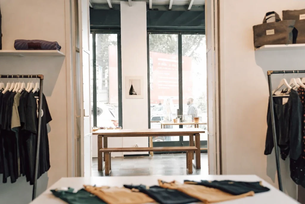 A minimalist clothing boutique interior with neatly folded shirts on a table, wooden bench seating, and large windows letting in natural daylight. Fashoin Product Development for an Apparel racks with dark and neutral clothing line both sides of the store, creating a clean retail display layout.