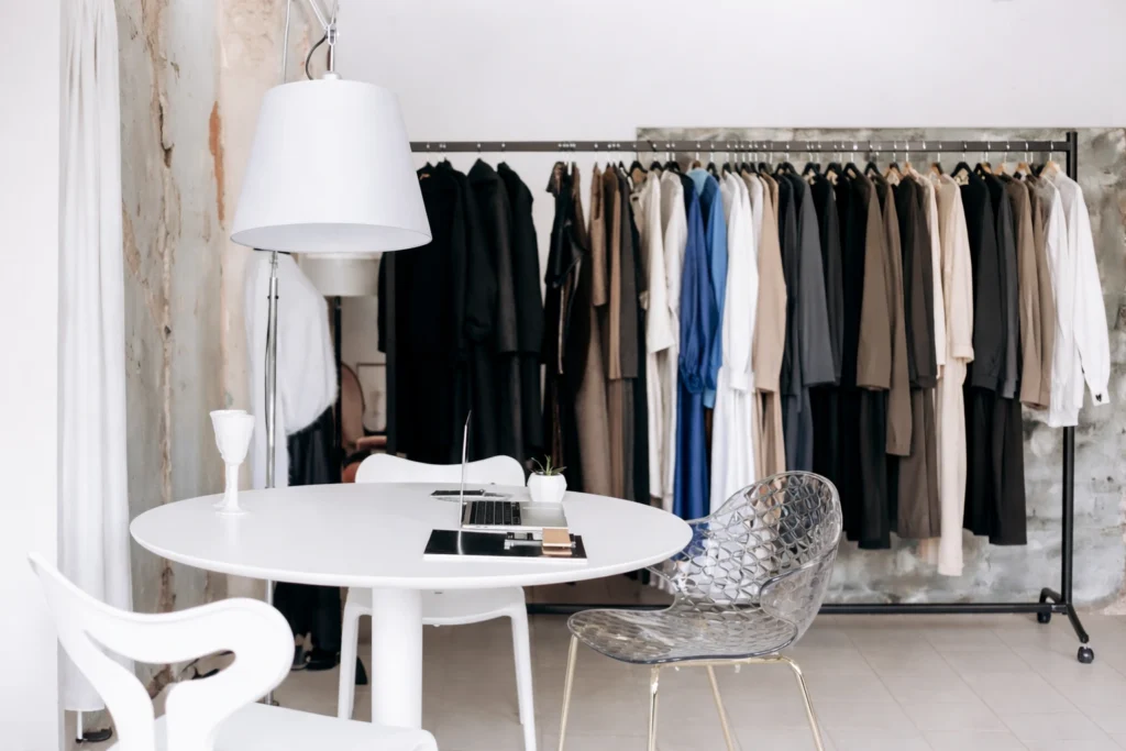 Contemporary fashion studio interior with a white round table, laptop, designer chair, and a clothing rack filled with neutral outfits in the background. Bright minimalist workspace representing private label fashion planning and apparel merchandising.