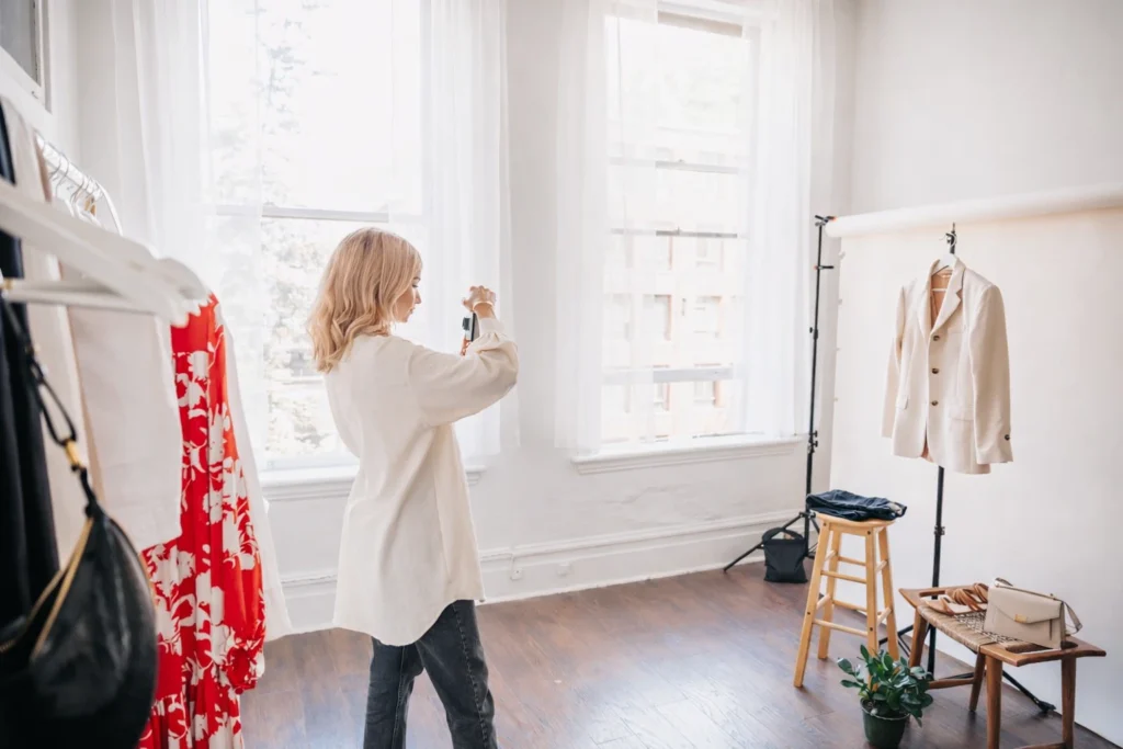 Blonde woman taking a photo of a beige blazer on a clothing stand inside a sunlit fashion studio with wooden floors and large windows. Modern fashion content creation scene highlighting apparel styling and private label strategy product photography.