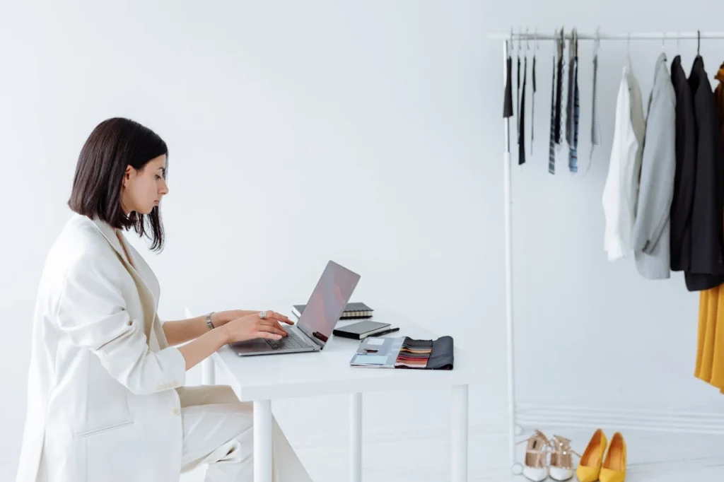 Fashion professional sitting at a white desk using a laptop with fabric swatches and garments hanging on a rack nearby in a bright studio. Private label strategy fashion planning and apparel design workspace with modern minimalist styling.